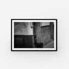 A black and white framed photograph of a man sitting down on a step of a house with a mask on.