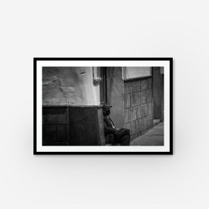 A black and white framed photograph of a man sitting down on a step of a house with a mask on.