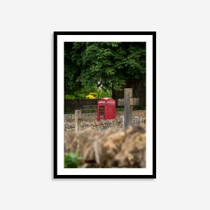A red phone box stands beside an old gravestone and a Christian cross in a Cotswolds village