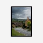 A moody photograph of a typical Cotswolds village with a road leading to traditional stone houses and lush green trees under a dramatic, cloudy sky.