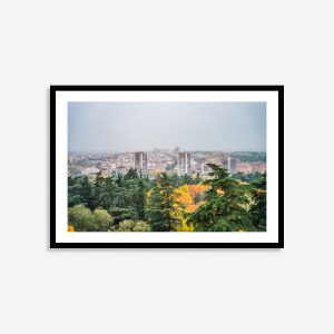 A sun-drenched viewpoint in Madrid, Spain, in autumn. The street is lined with trees displaying vibrant orange, green and yellow fall foliage against white buildings and blue sky.